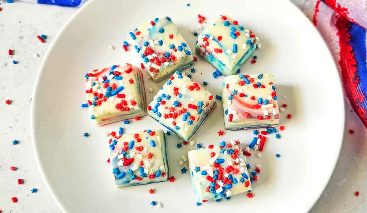 A white plate holds seven pieces of fudge decorated with red, white, and blue sprinkles, situated on a white surface with a red, white, and blue cloth nearby.