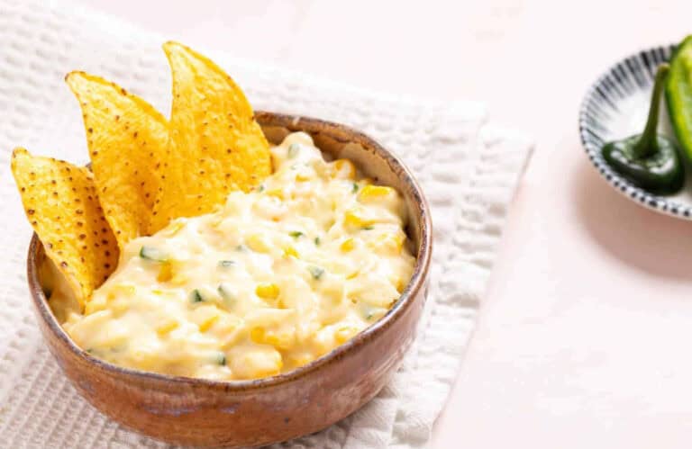 A bowl of creamy corn dip with chopped green herbs, served with three tortilla chips sticking out, placed on a white textured cloth. A plate with sliced jalapeños is in the background.