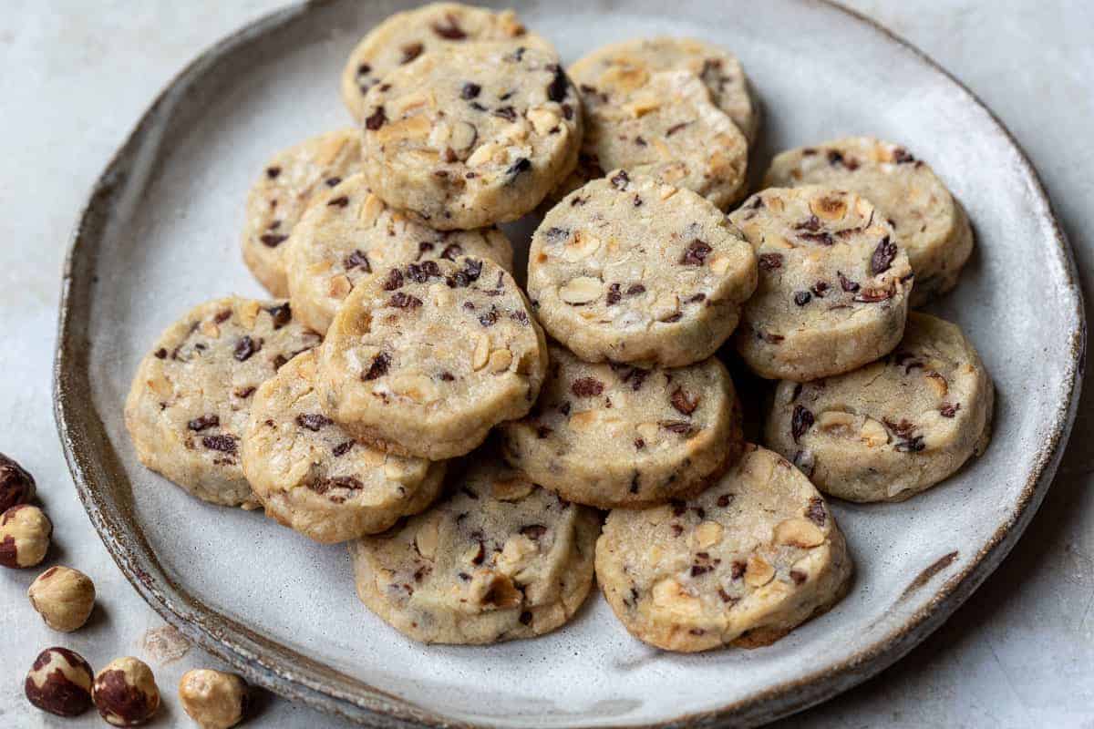 A ceramic plate holds a pile of round cookies with visible pieces of nuts and dried fruit; some hazelnuts rest beside the plate.