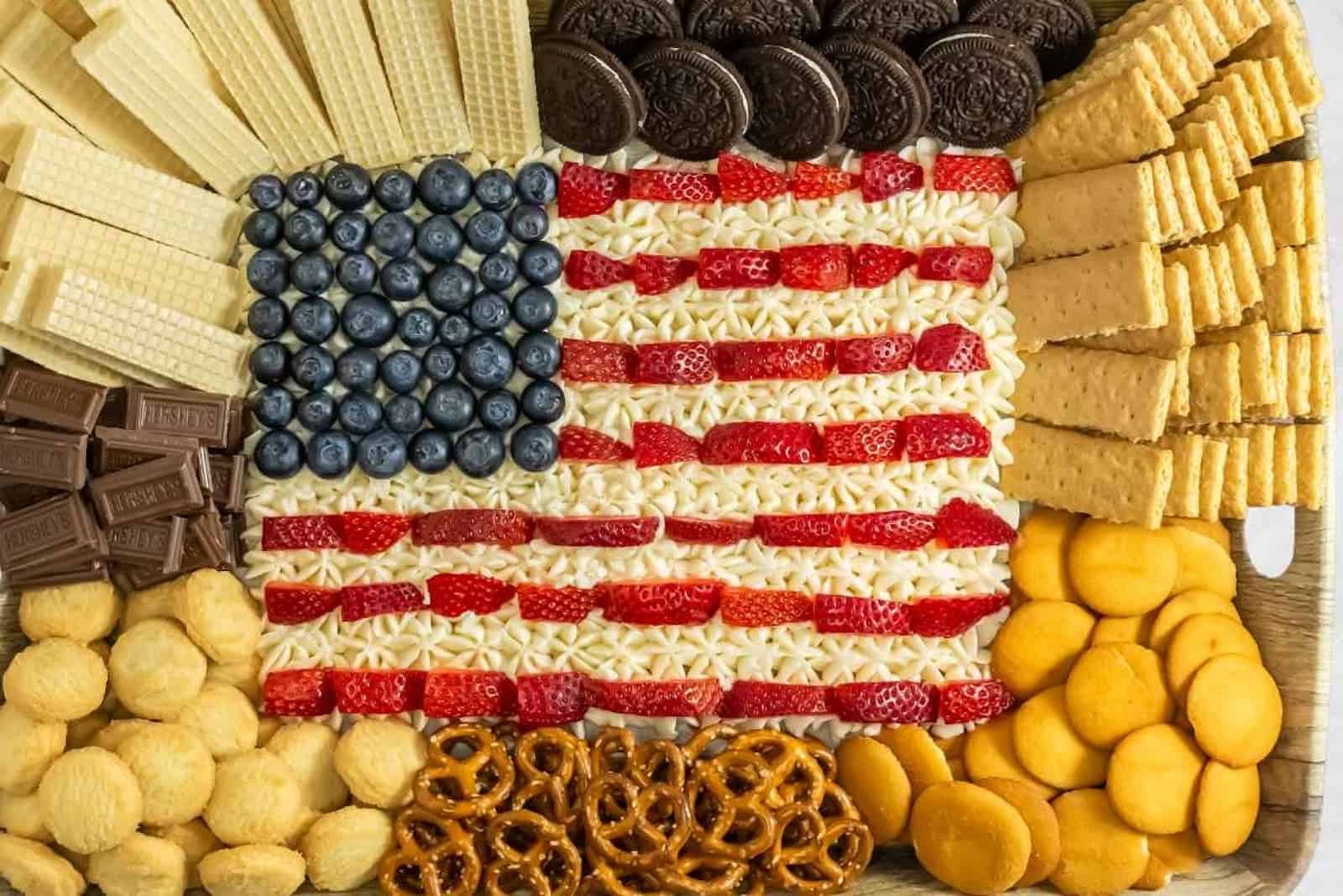 An assortment of snacks is arranged to resemble the American flag, including blueberries, strawberries, cream, wafers, cookies, pretzels, graham crackers, and chocolate pieces.