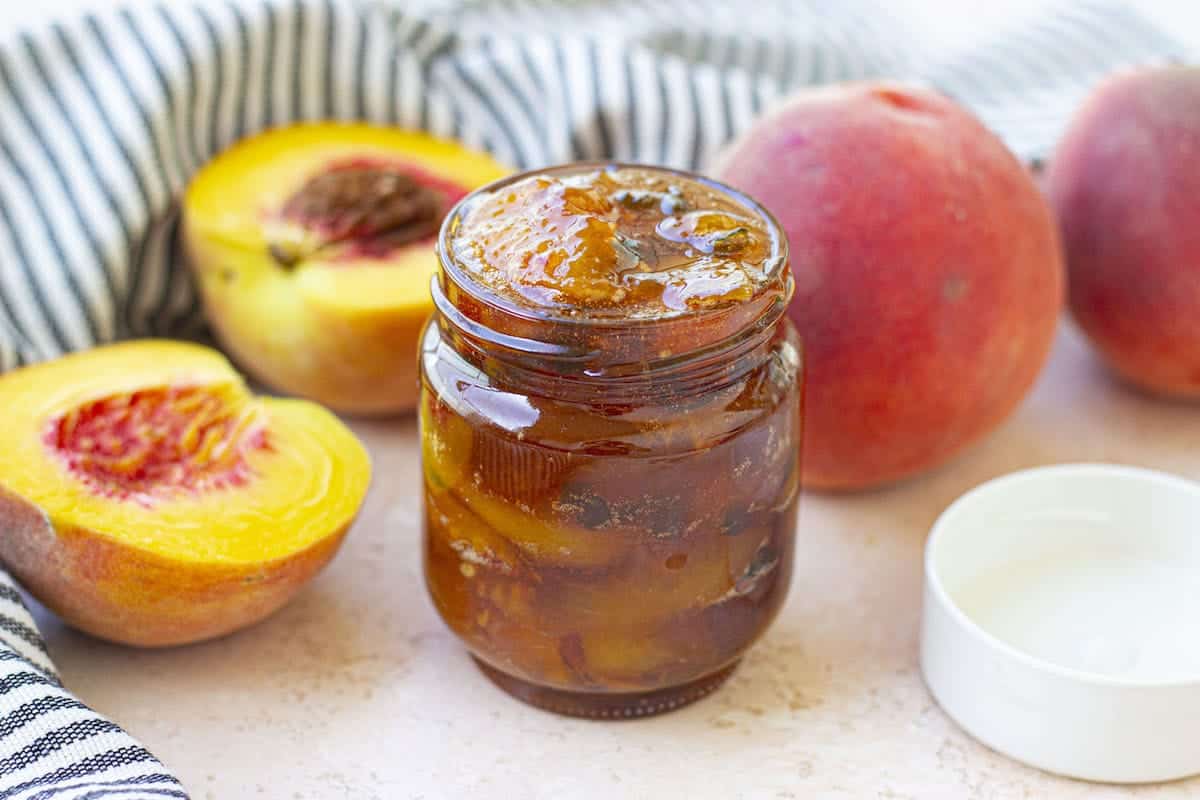 A glass jar of peach jam sits open on a counter beside fresh peaches, a striped cloth, and a white lid.