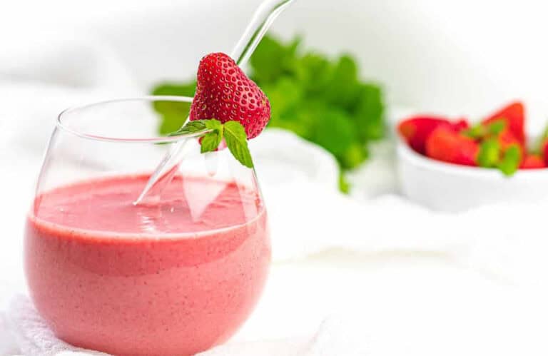 A glass of pink strawberry smoothie with a glass straw, garnished with a fresh strawberry. A bowl of strawberries is blurred in the background.