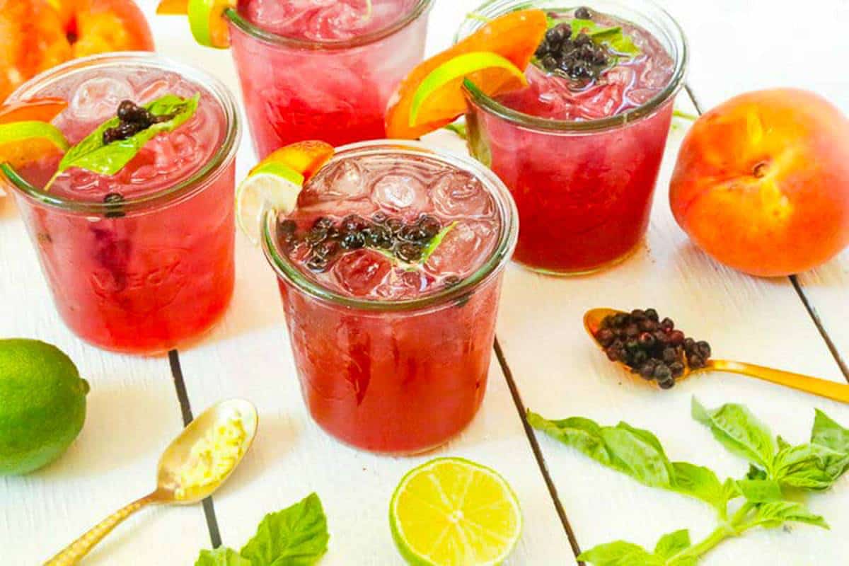 Four glasses of red iced drinks garnished with lime, peach slices, basil, and berries on a white table with fresh lime, peach, basil leaves, and a spoon nearby.