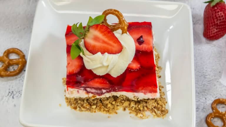 A slice of strawberry pretzel dessert topped with whipped cream, a strawberry half, and a pretzel on a white plate.