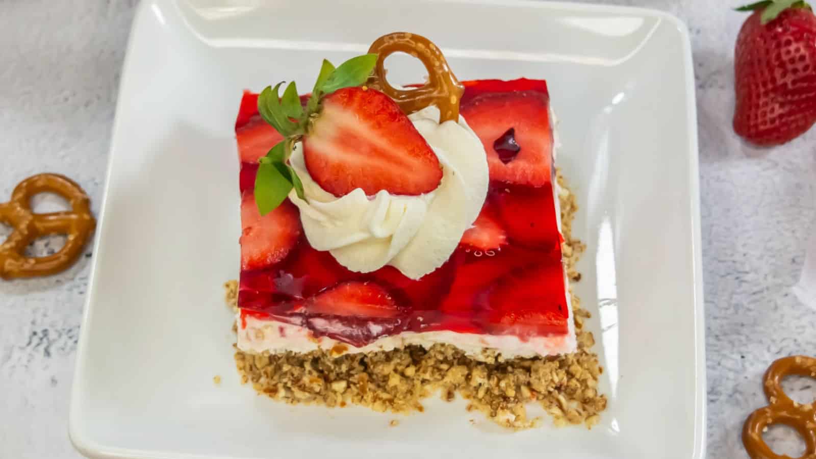 A slice of strawberry pretzel dessert topped with whipped cream, a strawberry half, and a pretzel on a white plate.