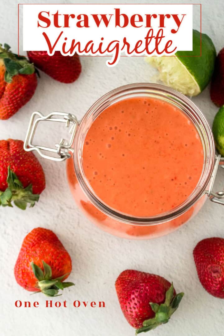 A jar of strawberry vinaigrette surrounded by fresh strawberries and a halved lime on a white surface.
