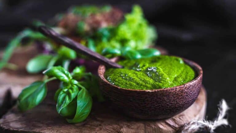 A wooden bowl filled with green pesto sauce, garnished with fresh basil leaves, sits on a rustic wooden surface.