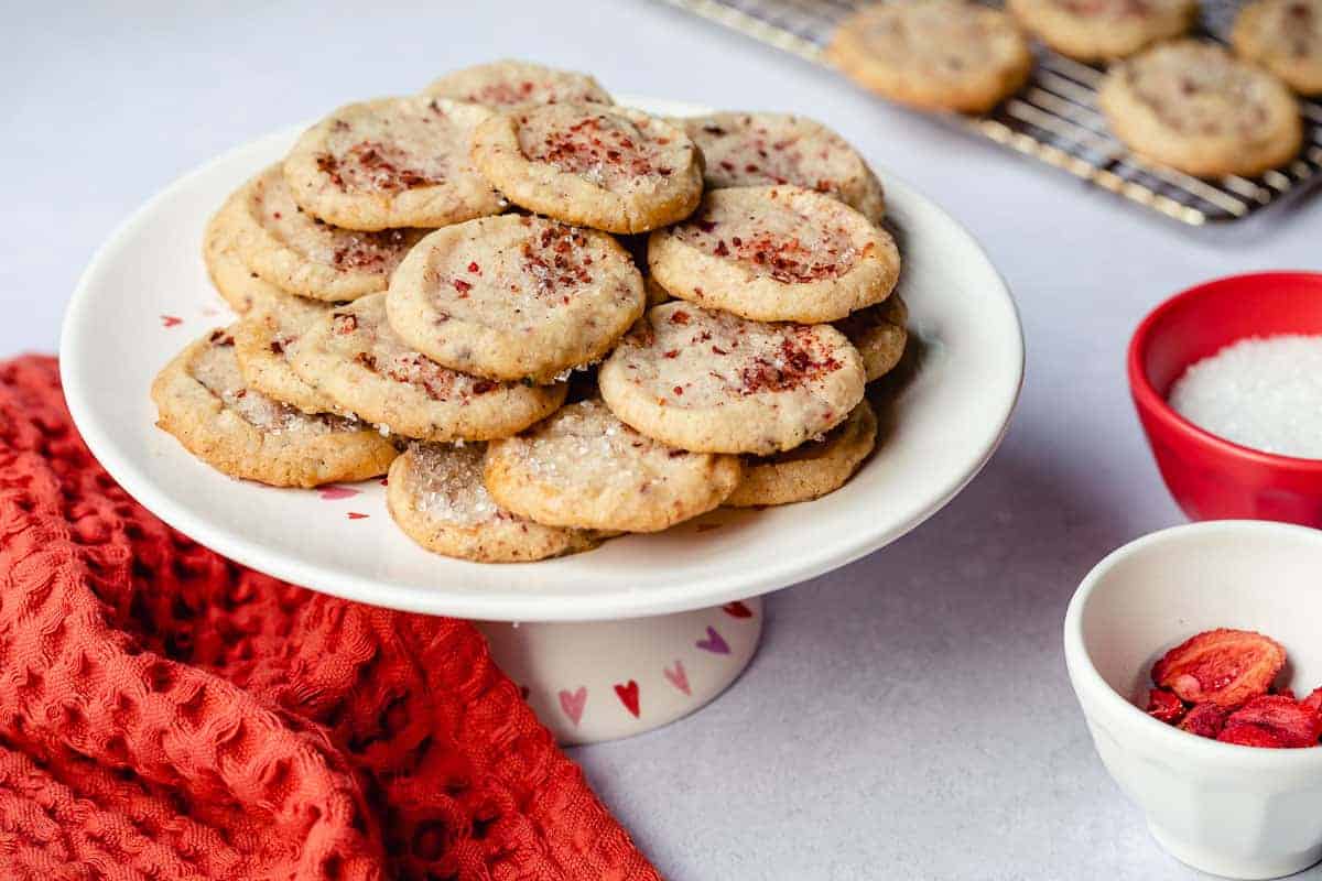A white cake stand holds a stack of cookies topped with red sprinkles. A cooling rack, red towel, and small bowls with sugar and dried fruit are nearby.