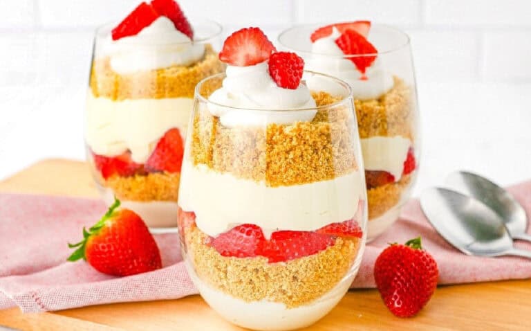 Three layered strawberry cheesecake parfaits in clear glasses, garnished with whipped cream and strawberries, are placed on a wooden surface with a pink cloth and two spoons nearby.