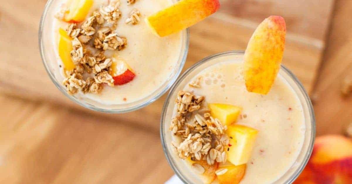 Two glasses of peach smoothie topped with granola and peach slices, placed on a wooden surface.