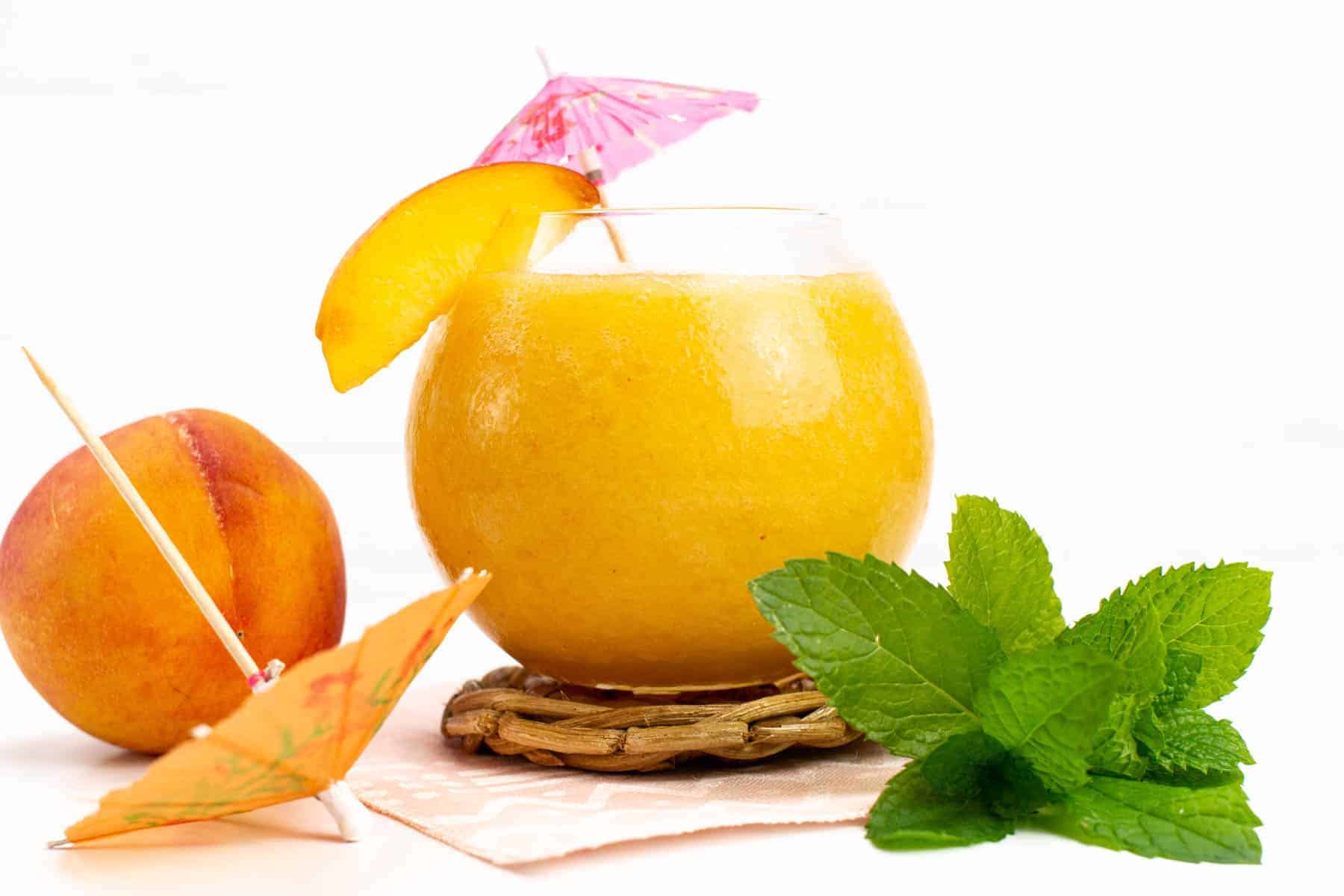 A glass of peach smoothie garnished with a peach slice and umbrella, next to a whole peach, fresh mint, and a cocktail umbrella on a white surface.