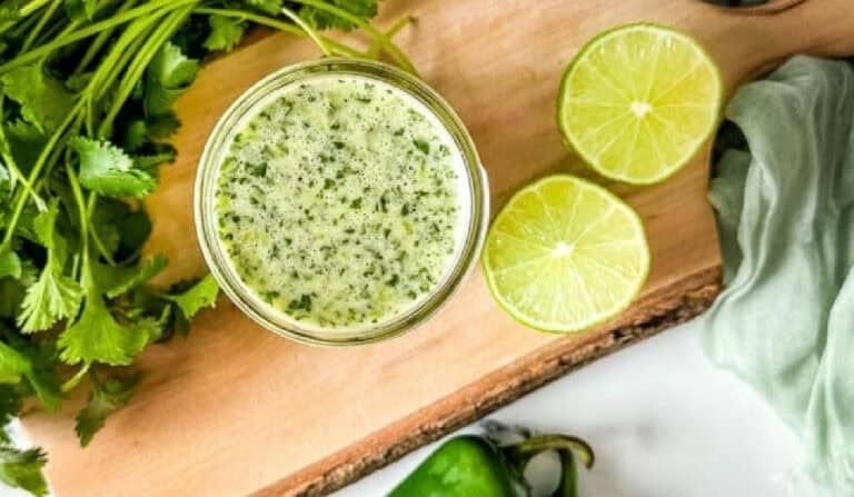 A glass jar filled with green sauce sits on a wooden board next to fresh cilantro, two lime halves, and a jalapeño pepper.