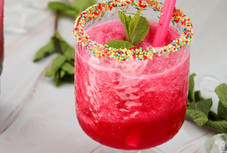 A glass of bright red frozen drink with a pink straw, topped with mint leaves and a rim coated in colorful sprinkles.
