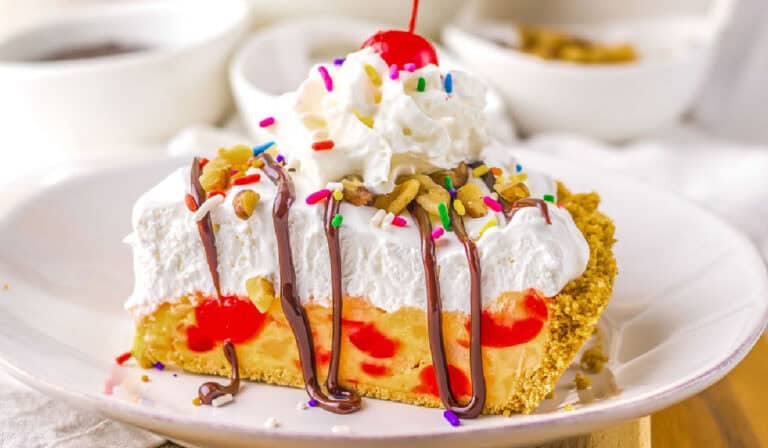 A slice of pie with whipped cream, chocolate drizzle, sprinkles, chopped nuts, and a cherry on top, served on a white plate.