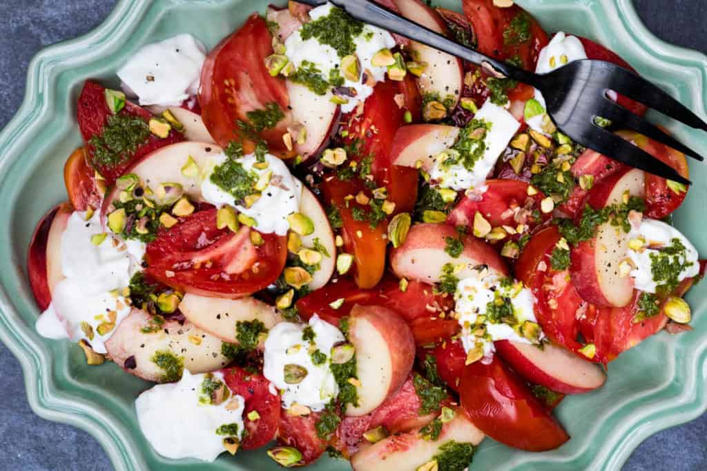 A salad with sliced tomatoes, peaches, dollops of cheese, chopped pistachios, and green herb dressing served on a green platter with a black serving fork.