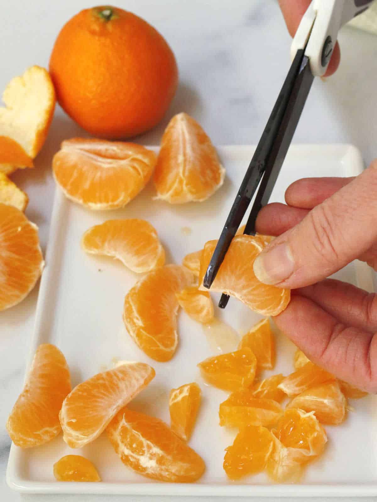 A person uses scissors to cut an orange segment in half on a white plate with peeled orange segments and a whole orange nearby.