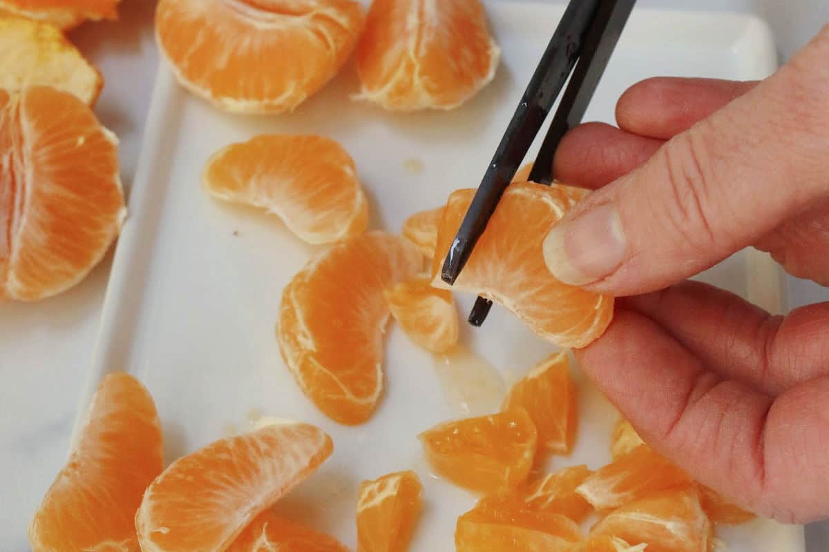 A hand uses scissors to cut the white membrane off a tangerine segment, with other peeled tangerine pieces on a plate nearby.