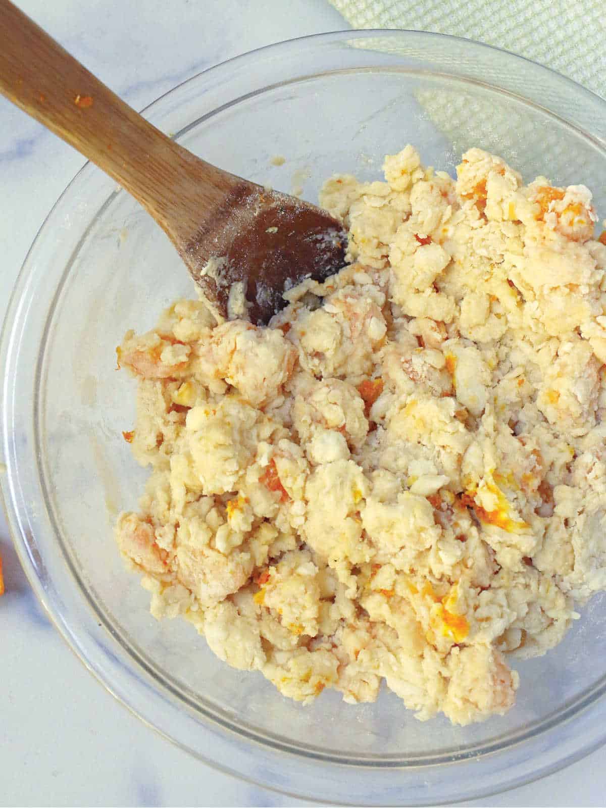 A glass bowl containing partially mixed biscuit dough with orange flecks, being stirred with a wooden spoon.