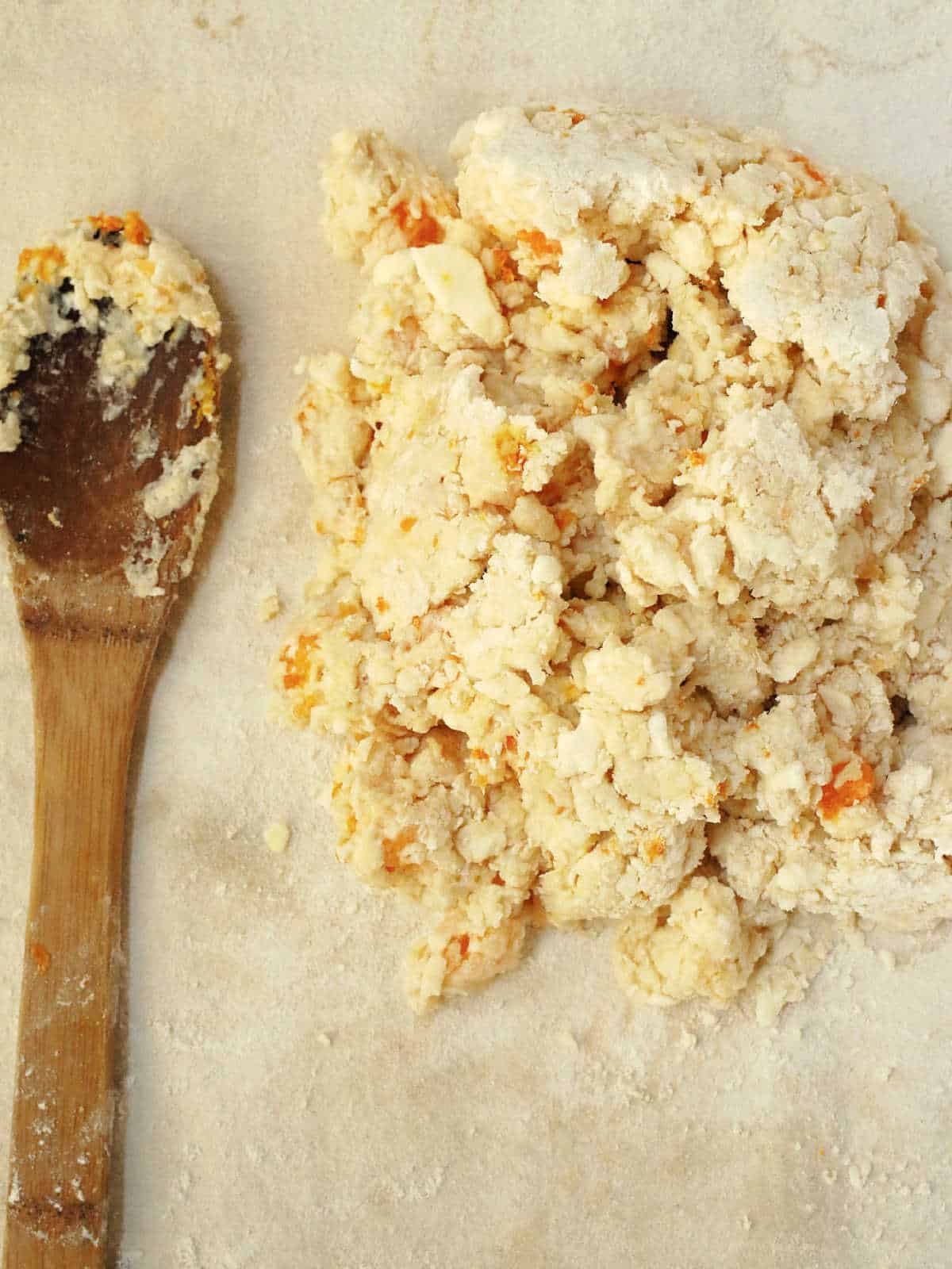 A wooden spoon lies next to a mound of rough, crumbly scone dough on a floured surface.