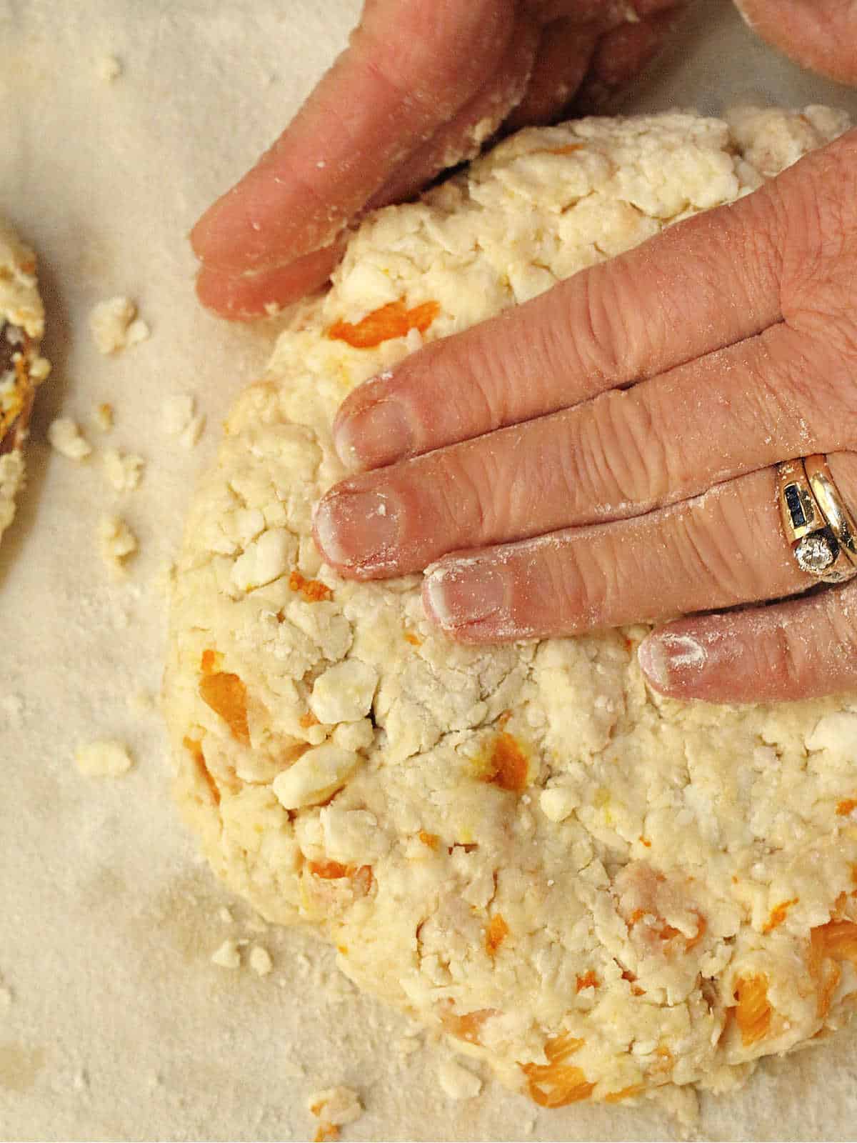 Close-up of hands pressing and shaping scone dough with visible chunks of fruit on a floured surface.