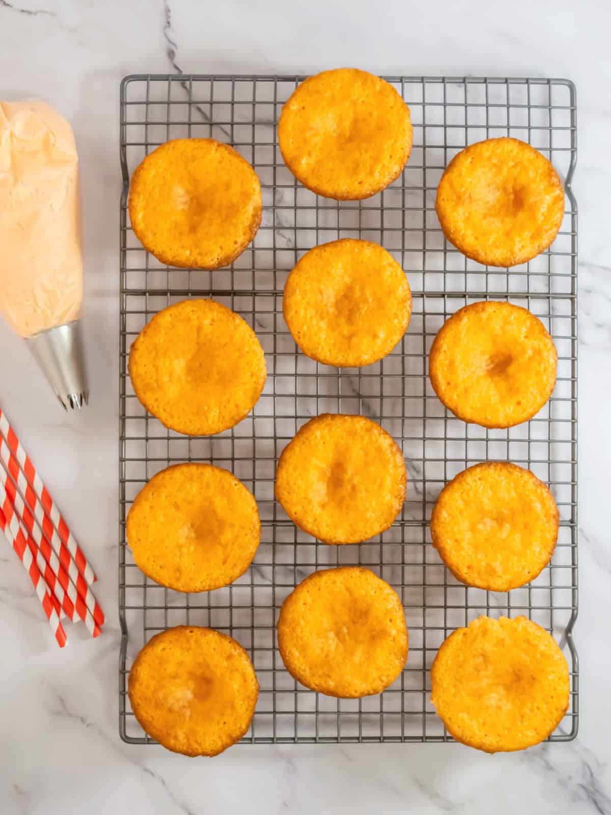 Twelve unfrosted cupcakes arranged on a cooling rack, with a piping bag of orange frosting and striped straws beside them on a marble surface.