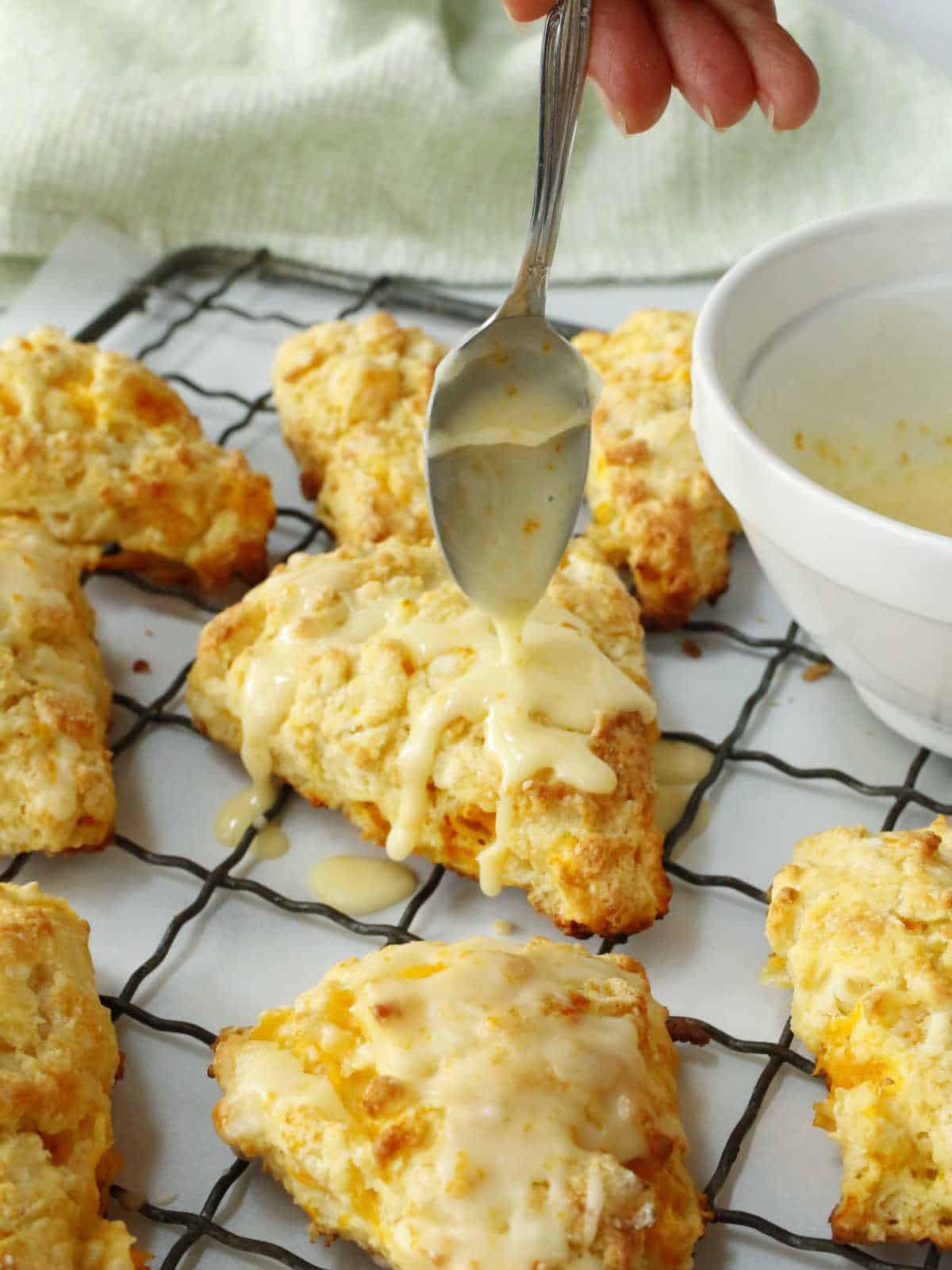 Drizzling a glaze from a spoon onto a citrus scone resting on a wire rack, with a white bowl of glaze nearby.