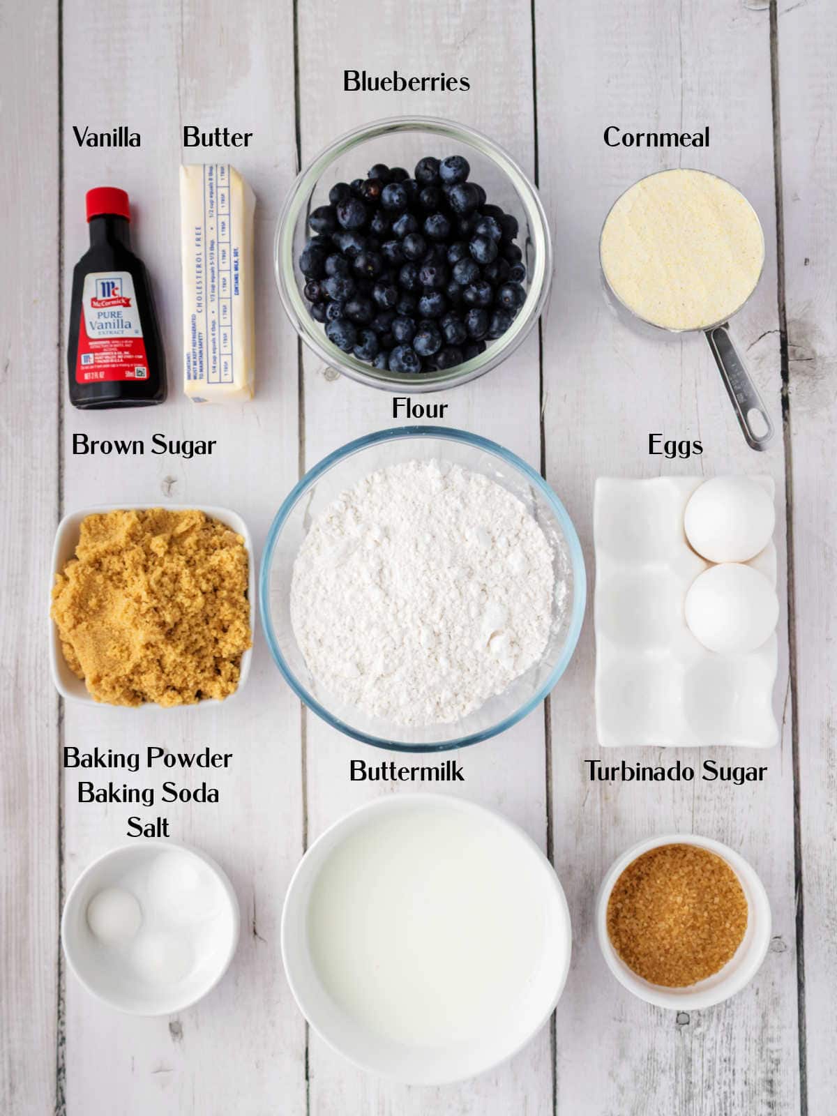 Various labeled baking ingredients, including blueberries, butter, flour, cornmeal, eggs, two types of sugar, buttermilk, vanilla, and a mixture of baking powder, baking soda, and salt.