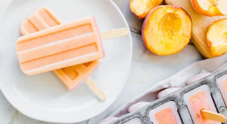 Two peach-colored popsicles on a white plate beside fresh peach halves and a tray of frozen popsicle molds.