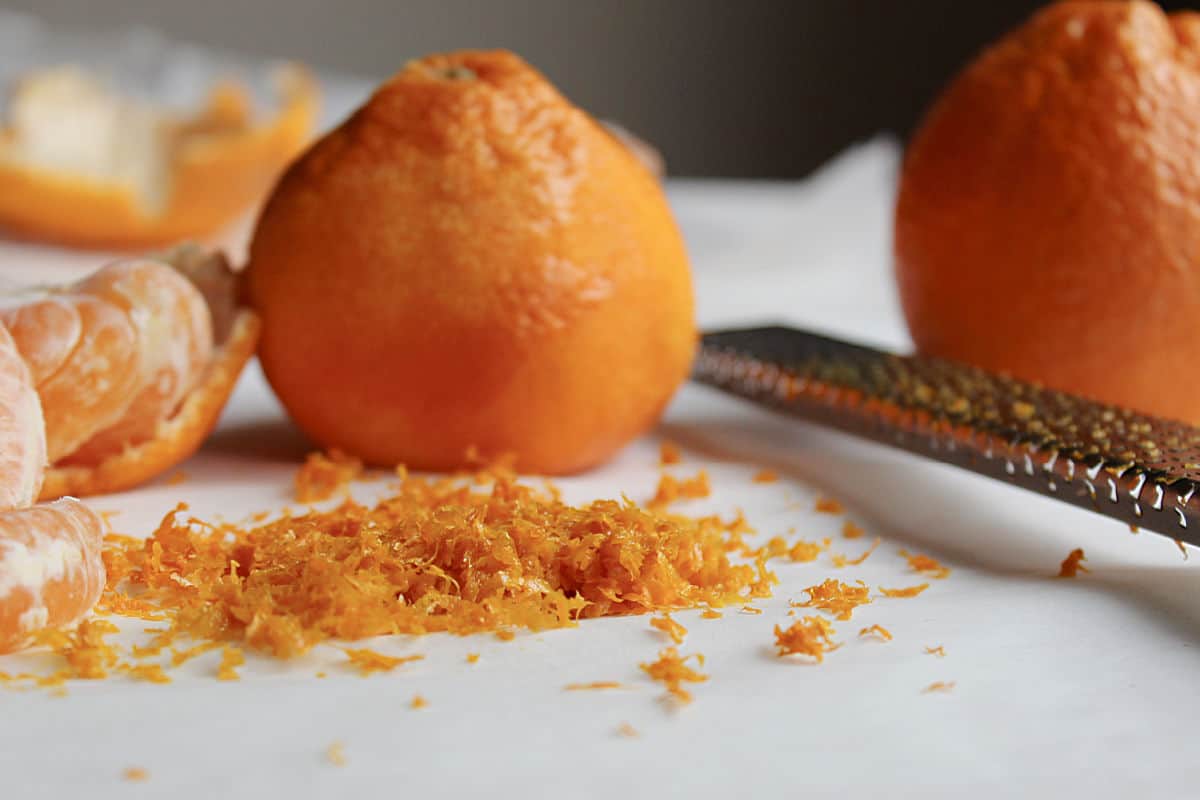 Whole and peeled oranges with a pile of orange zest and a metal grater on a white surface.