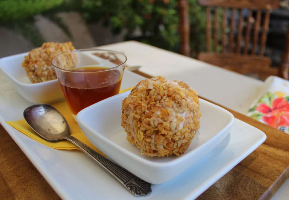 Two scoops of fried ice cream in white bowls with a cup of honey and a metal spoon on a rectangular white plate.