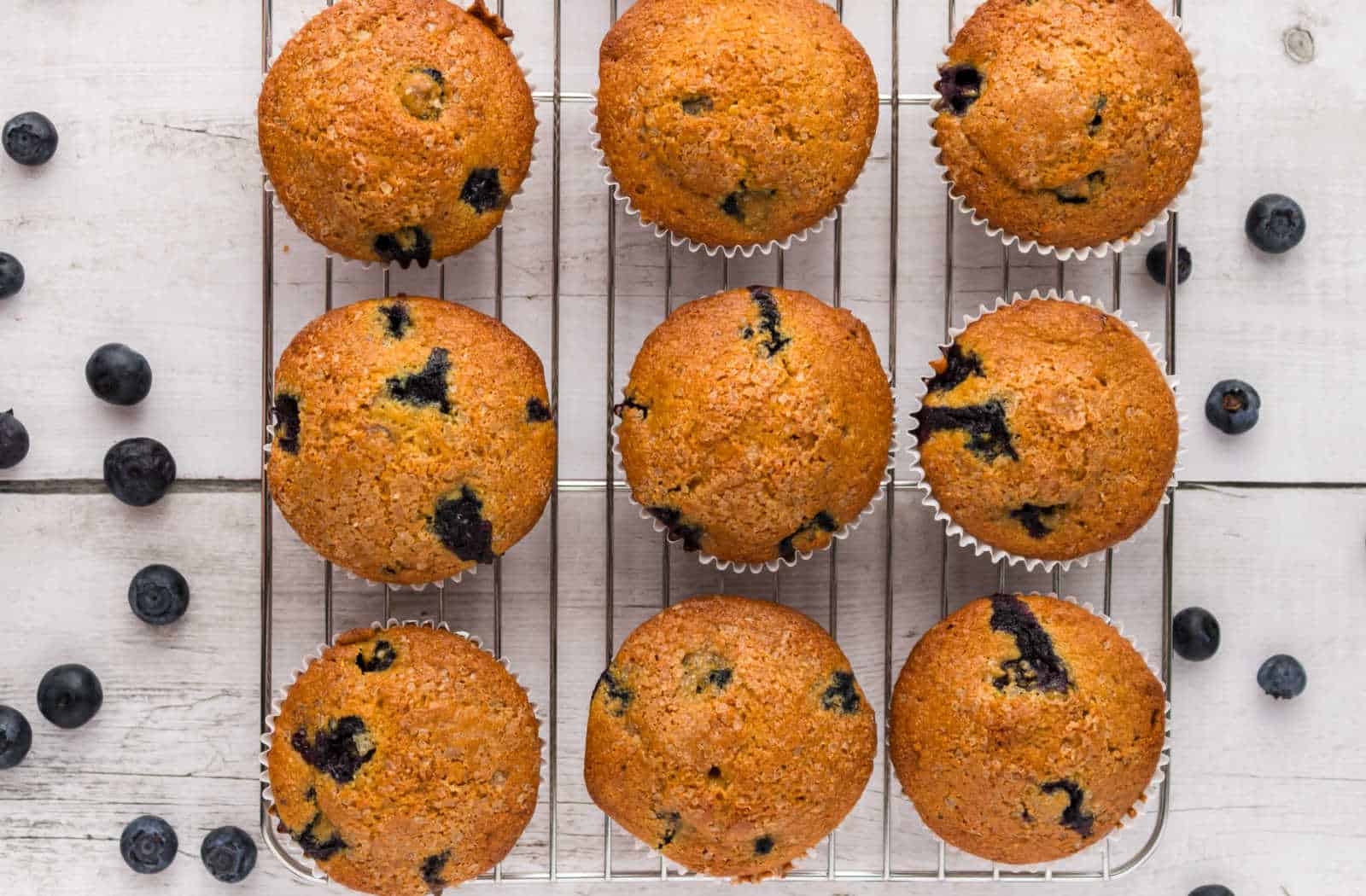 Nine blueberry muffins cooling on a wire rack, with scattered fresh blueberries on a white wooden surface.
