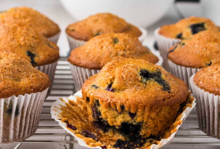 Blueberry muffins with golden tops cool on a wire rack, with one muffin partially unwrapped to show the berries inside.