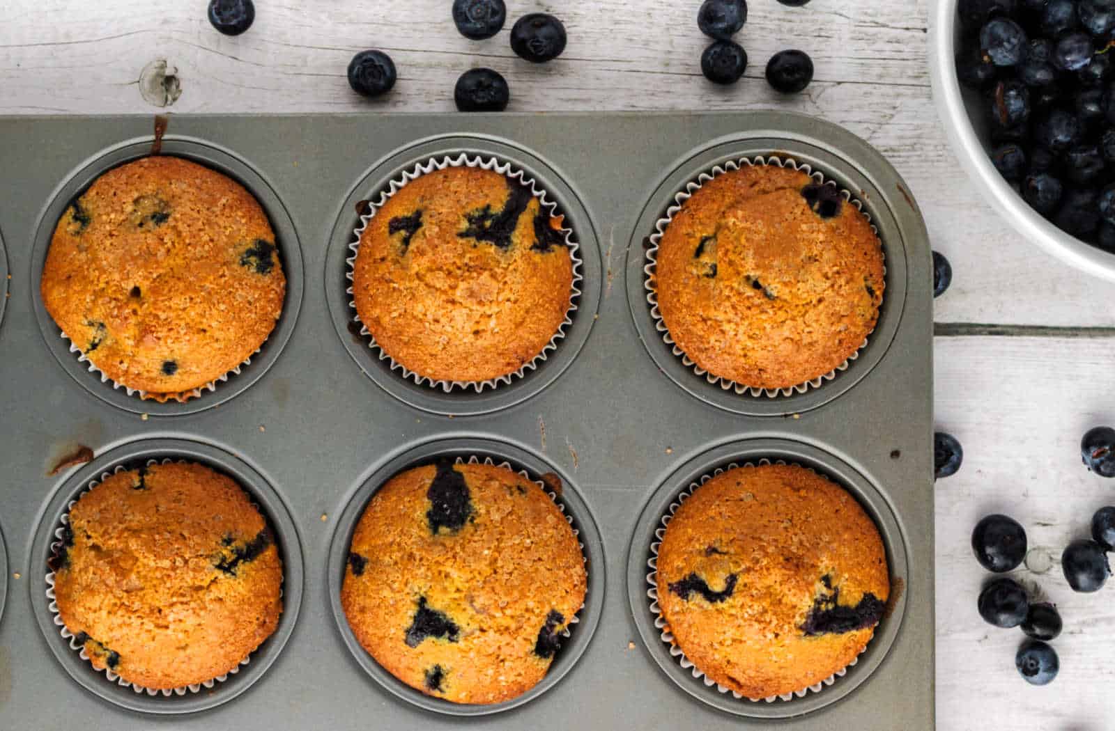 A muffin tin holding six baked blueberry muffins sits on a white wooden surface, with fresh blueberries scattered around and a bowl of blueberries nearby.