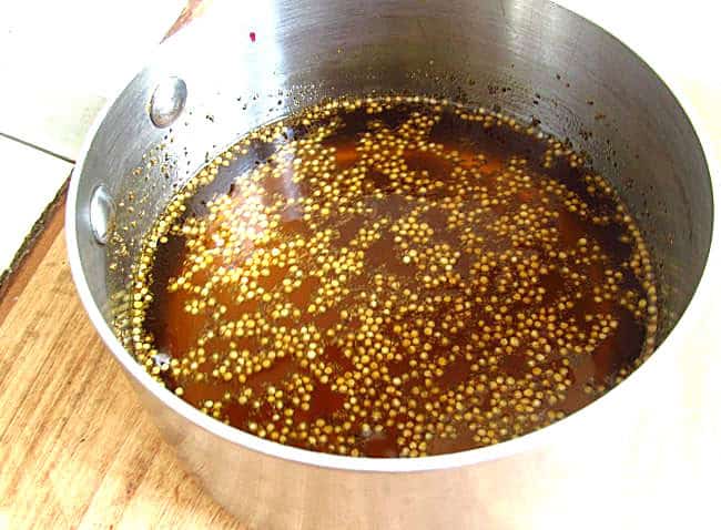 A stainless steel pot containing a brownish liquid with yellow mustard seeds floating on the surface, sitting on a wooden surface.
