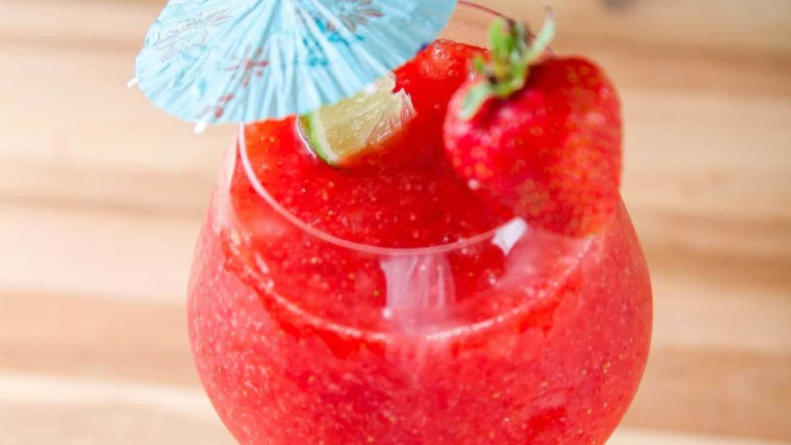 A red frozen drink in a glass garnished with a strawberry, lime wedge, and a blue cocktail umbrella, on a wooden surface.