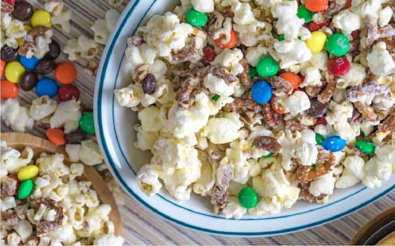 A bowl of popcorn mixed with colorful candy-coated chocolates and pretzel pieces on a striped surface, with extra pieces scattered around.