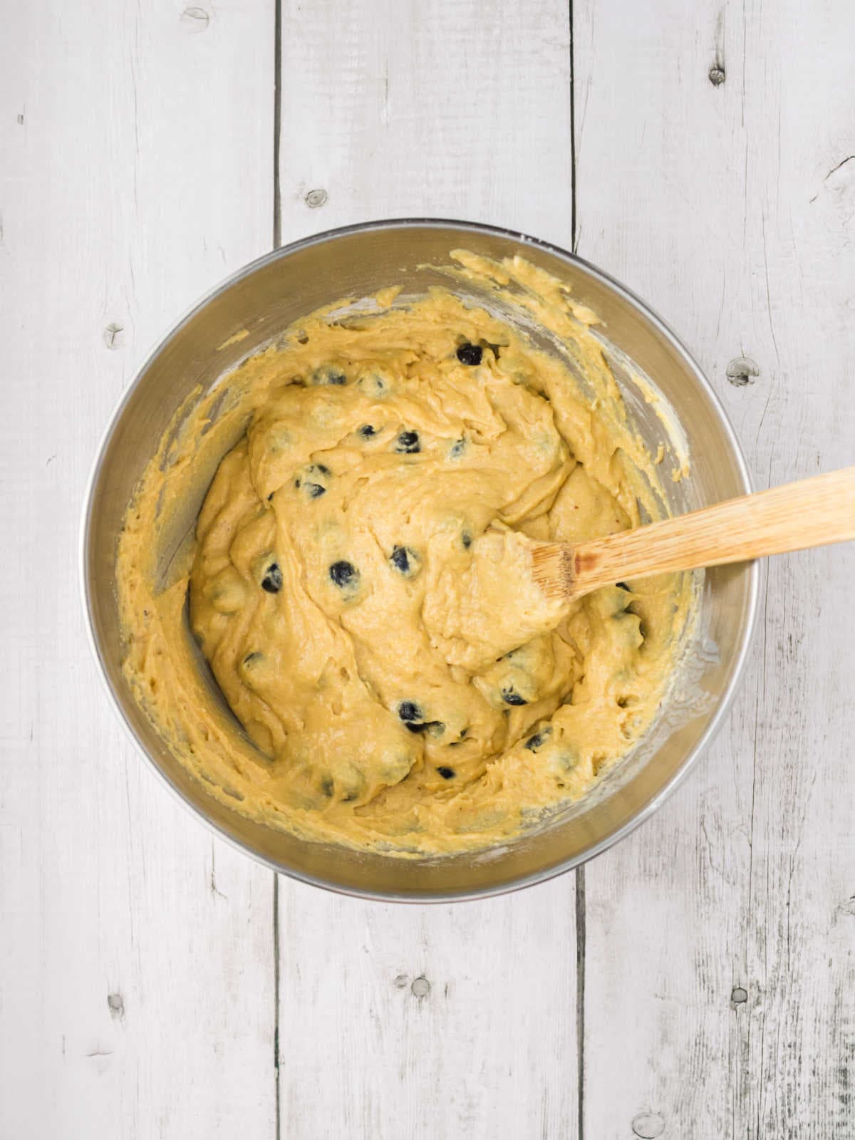 A metal mixing bowl filled with blueberry muffin batter and a wooden spatula, placed on a white wooden surface.