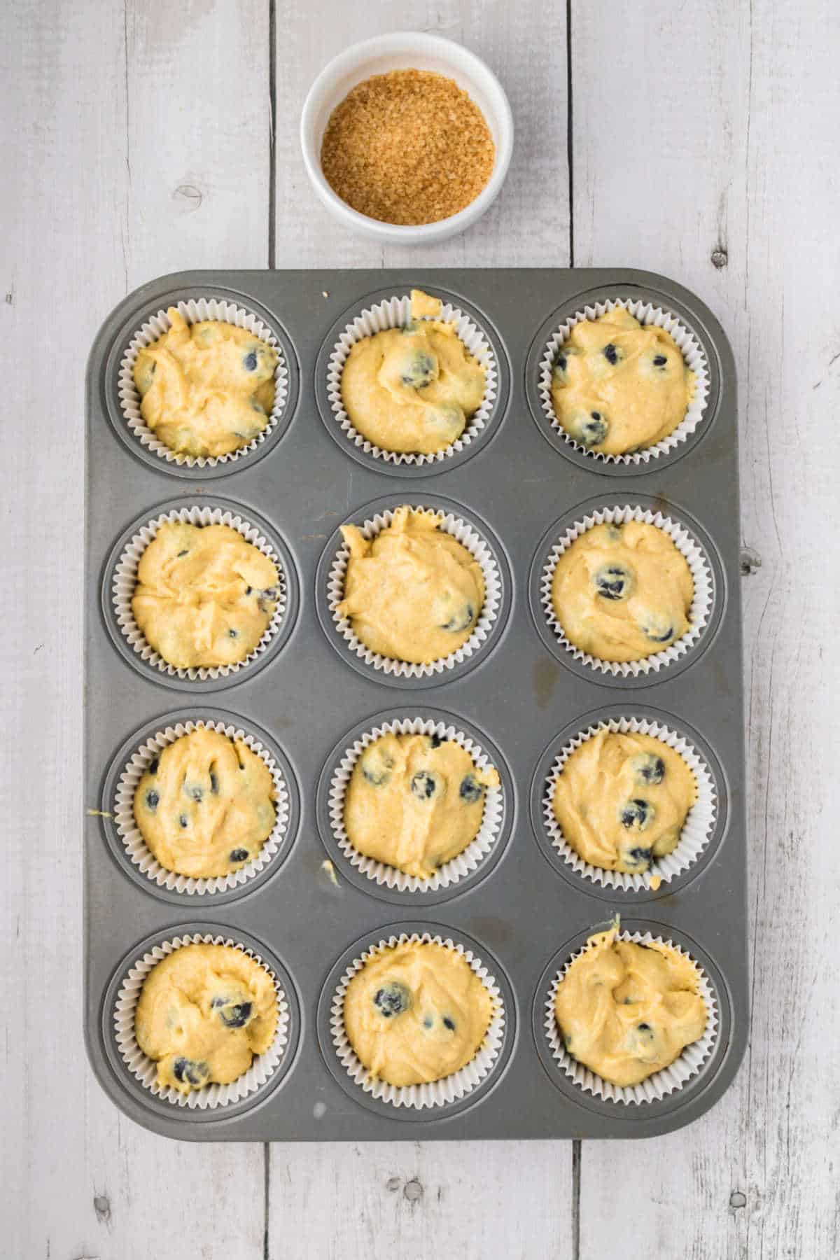 Muffin tin filled with blueberry muffin batter in paper liners, placed on a white wooden surface, with a small bowl of brown sugar above the tin.