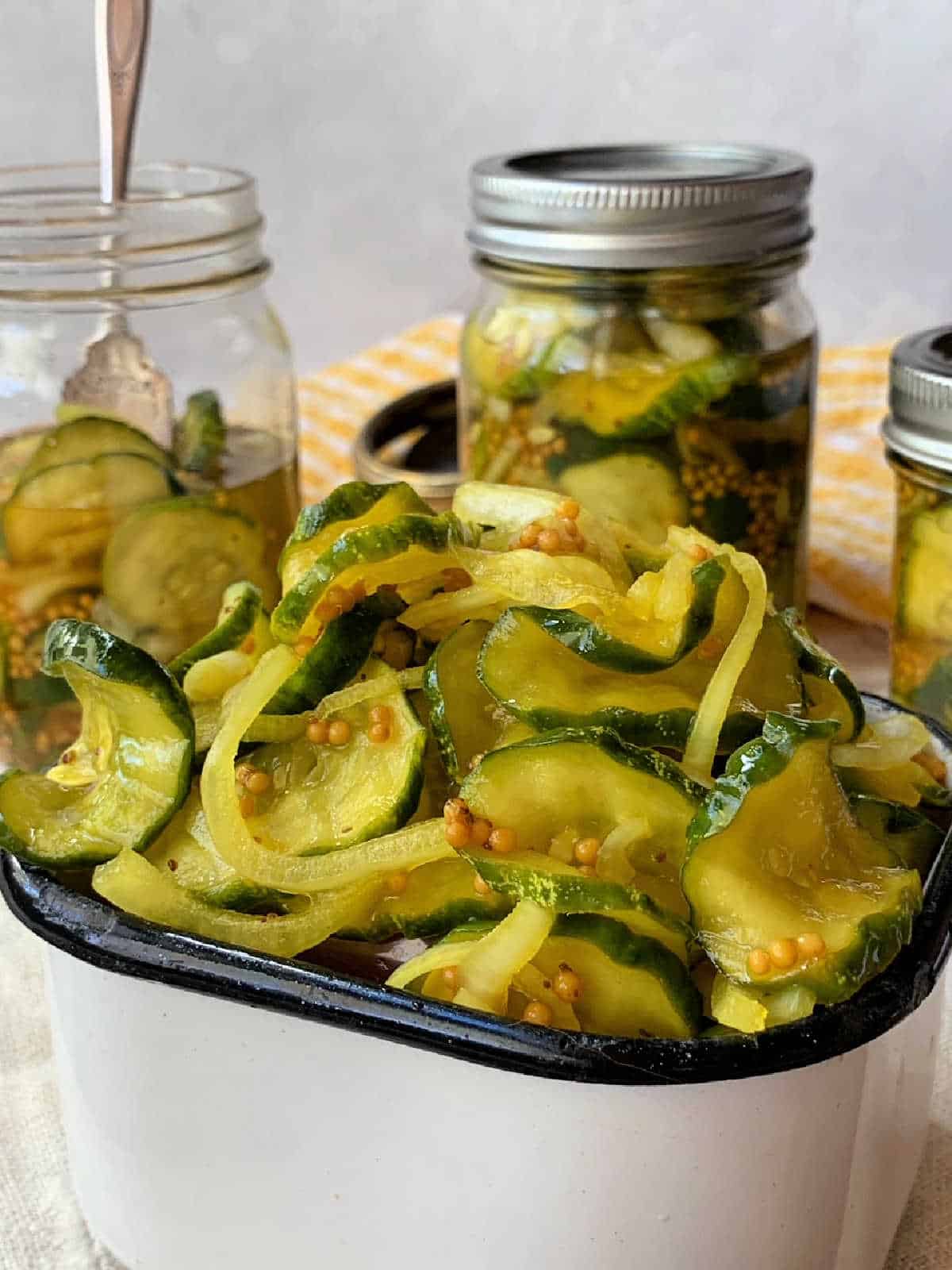 A white dish filled with sliced pickles sits in front of jars containing more pickles and brine, with a fork and yellow plaid cloth in the background.
