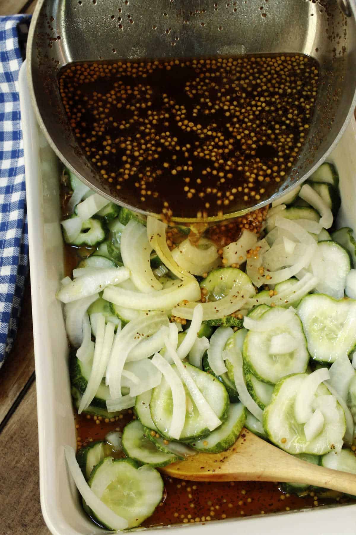 Sliced cucumbers and onions in a dish with a wooden spoon as a bowl of vinegar-based dressing with mustard seeds is being poured over them.