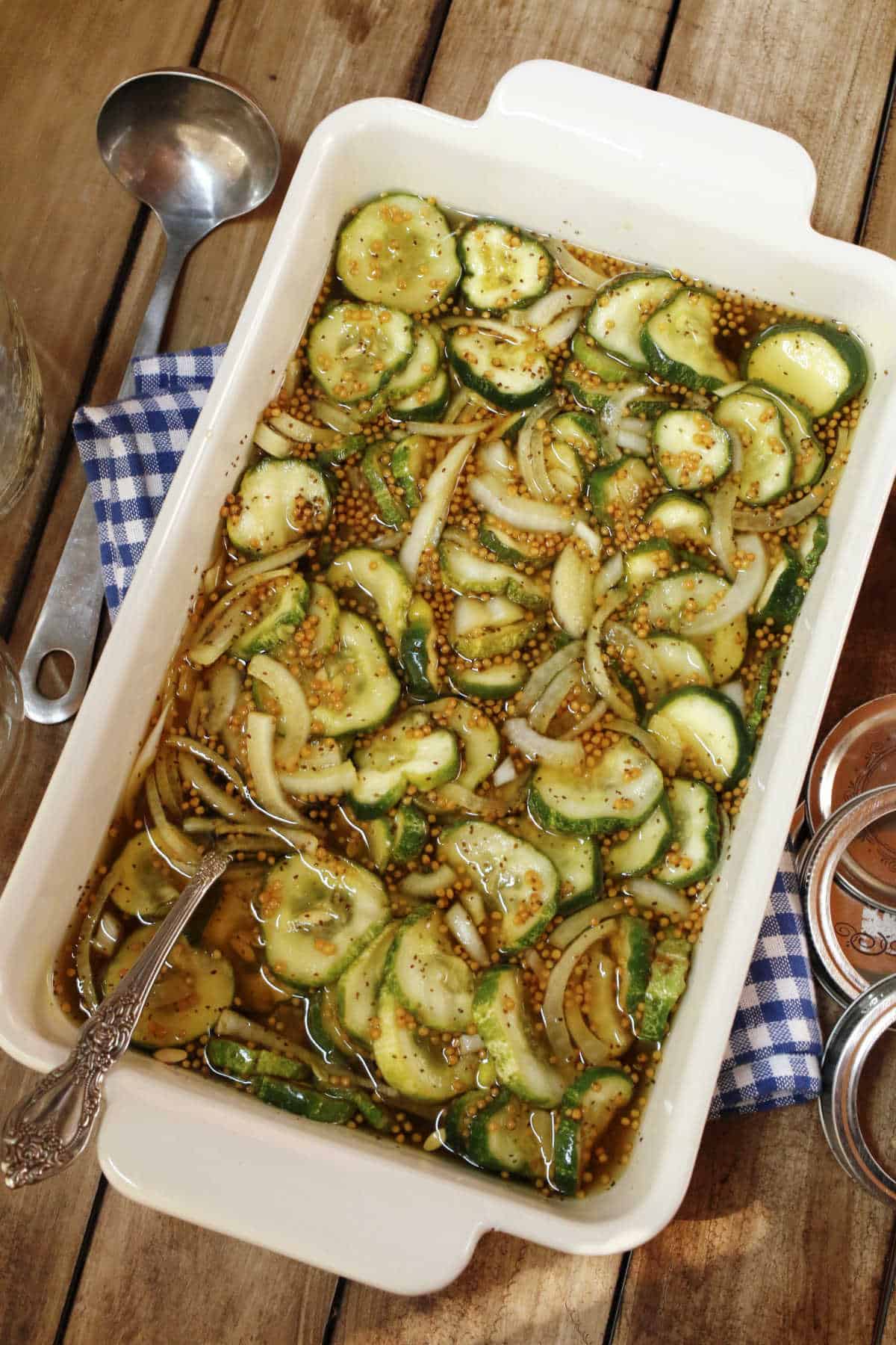 A rectangular dish filled with sliced cucumbers, onions, and pickling spices in brine, with a spoon and kitchen towel on a wooden table.