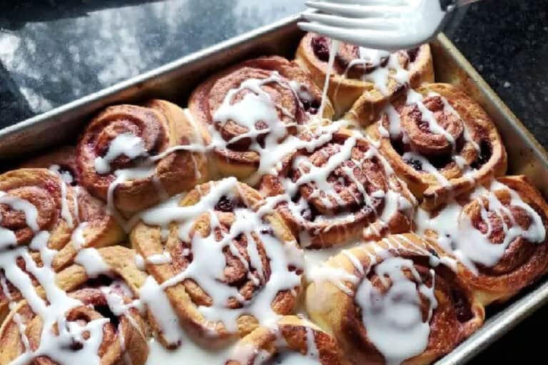 A tray of cinnamon rolls is being drizzled with white icing using a fork.