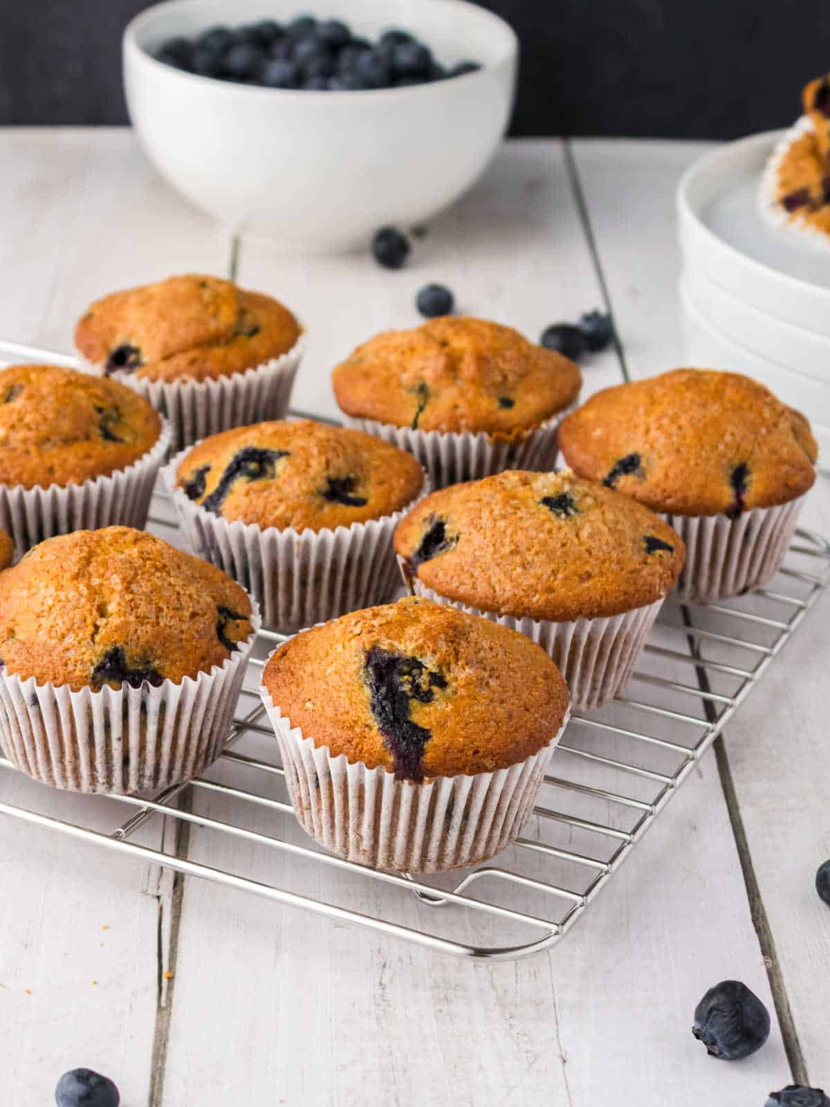 Nine blueberry muffins on a cooling rack with fresh blueberries and a bowl of blueberries in the background on a white wooden surface.