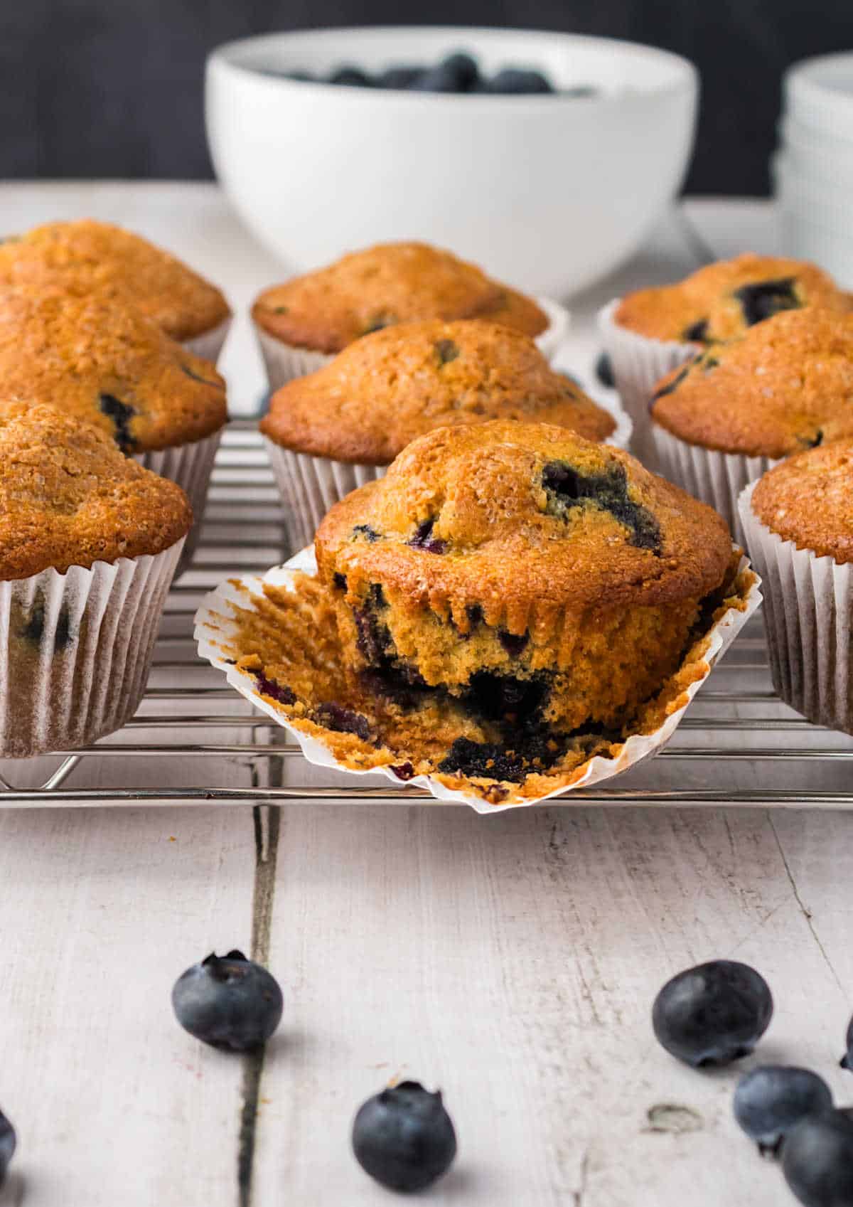 A batch of blueberry cornmeal muffins on a cooling rack, with one muffin unwrapped to show the blueberries inside; scattered blueberries and a bowl are in the background.