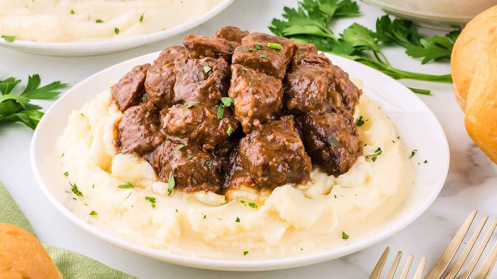 A plate of mashed potatoes topped with chunks of beef in brown gravy, garnished with chopped herbs, with parsley and a roll on the side.