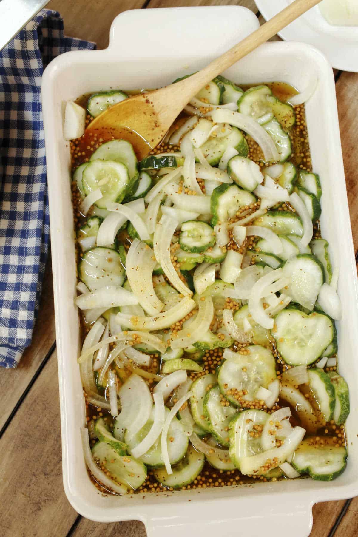A white baking dish filled with sliced cucumbers, onions, and mustard seeds in a vinaigrette marinade, with a wooden spoon and a blue checkered napkin nearby.