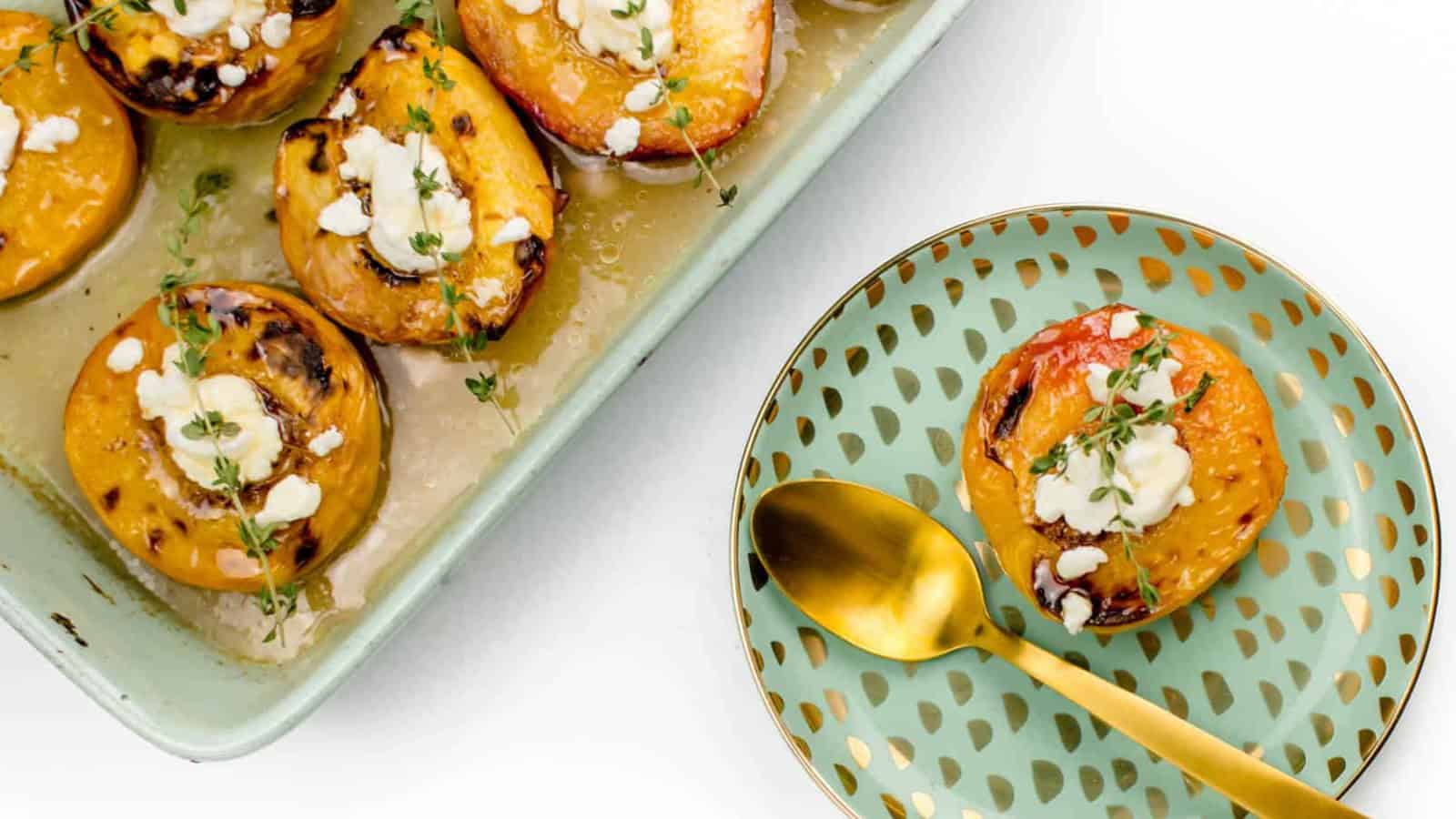 Halved roasted peaches topped with cheese and herbs are served on a decorative plate with a gold spoon, next to a baking dish containing more peaches.
