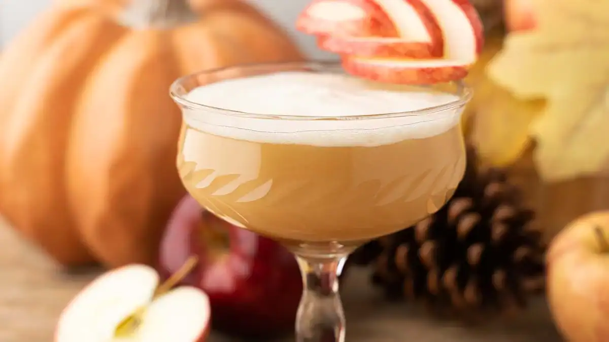 A frothy tan cocktail in a coupe glass garnished with apple slices, with pumpkins, apples, pinecones, and autumn leaves in the background.