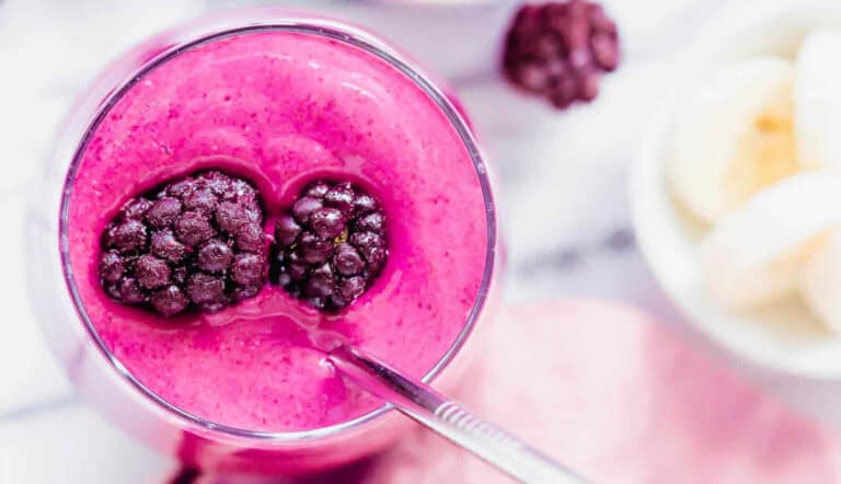 A glass of bright pink smoothie topped with two blackberries, with a metal straw inserted and a bowl of banana slices in the background.