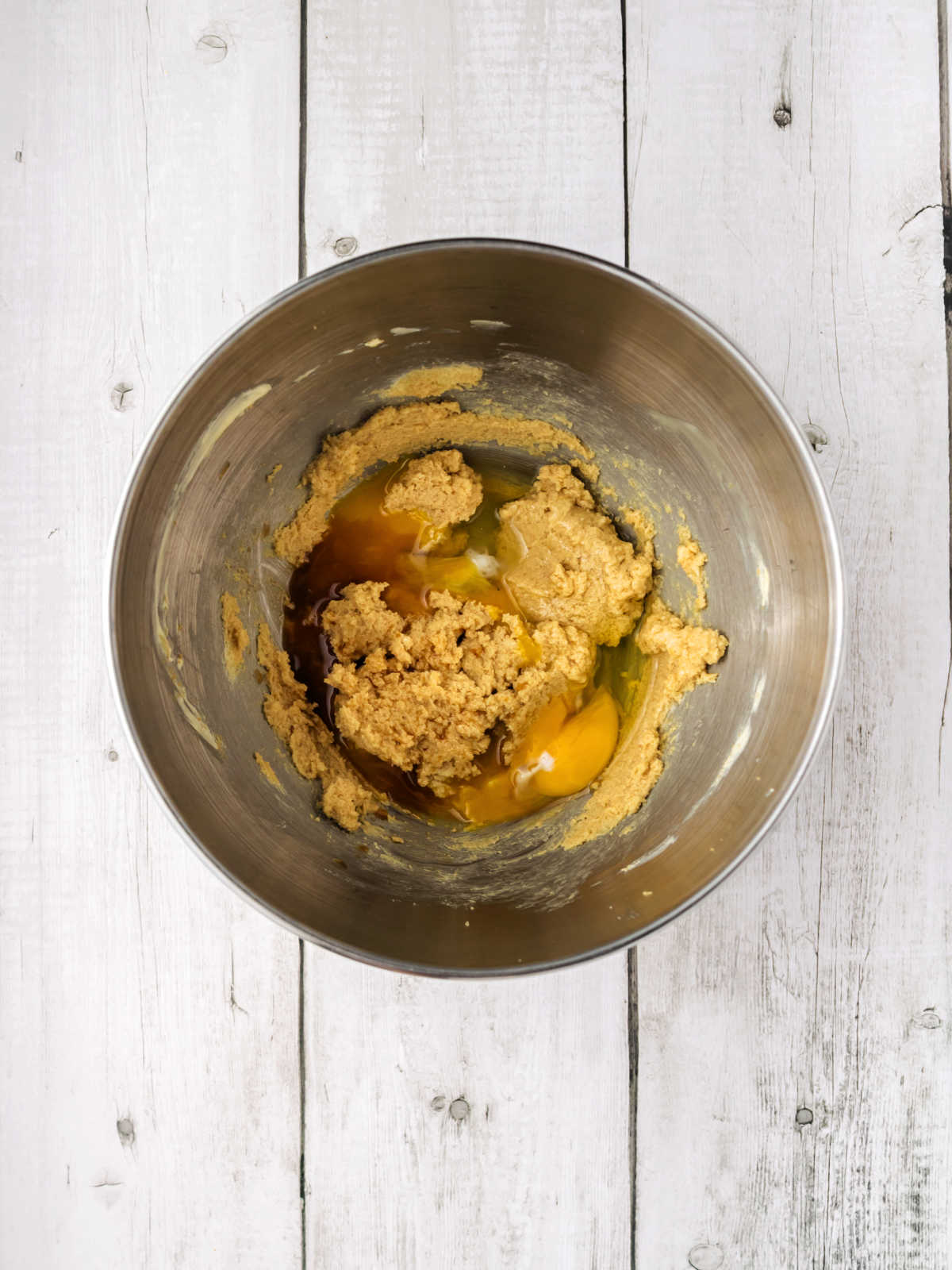 A metal mixing bowl containing creamed butter and sugar with two cracked eggs on top, placed on a white wooden surface.